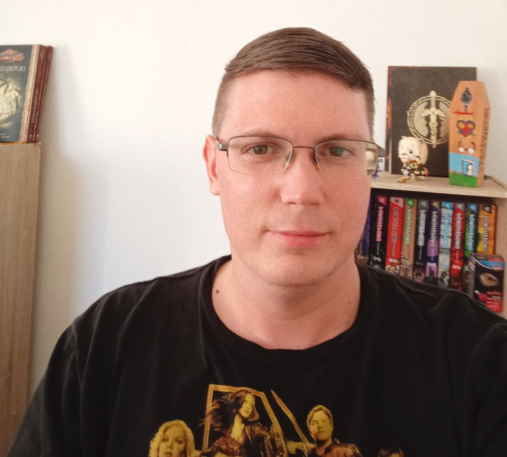 Simon Crain. Ein junger Mann mit  Site-Cutt Frisur, braune Haare, Brille und Sommersprossen, schaut lächelnd in die Kamera. hinter ihm ein Bücherschrank.