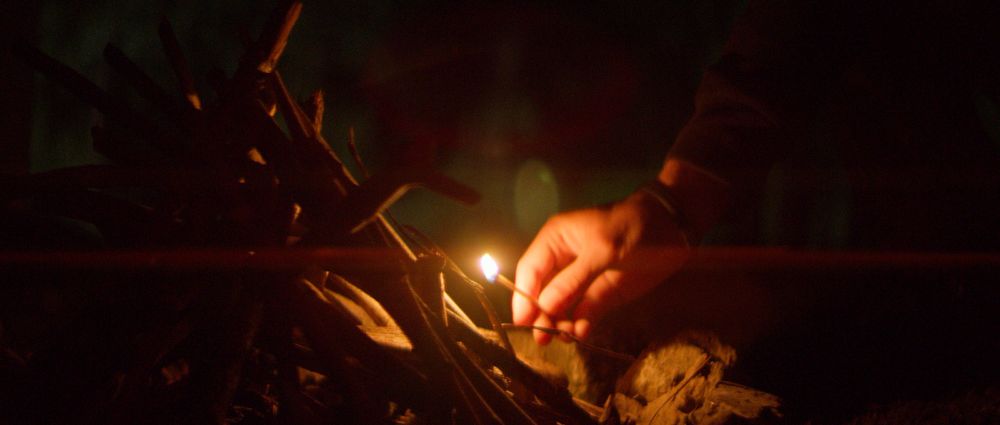 Image extrait d'un court métrage.     Gros plan de nuit sur une main tenant une allumette devant un petit tas de bois