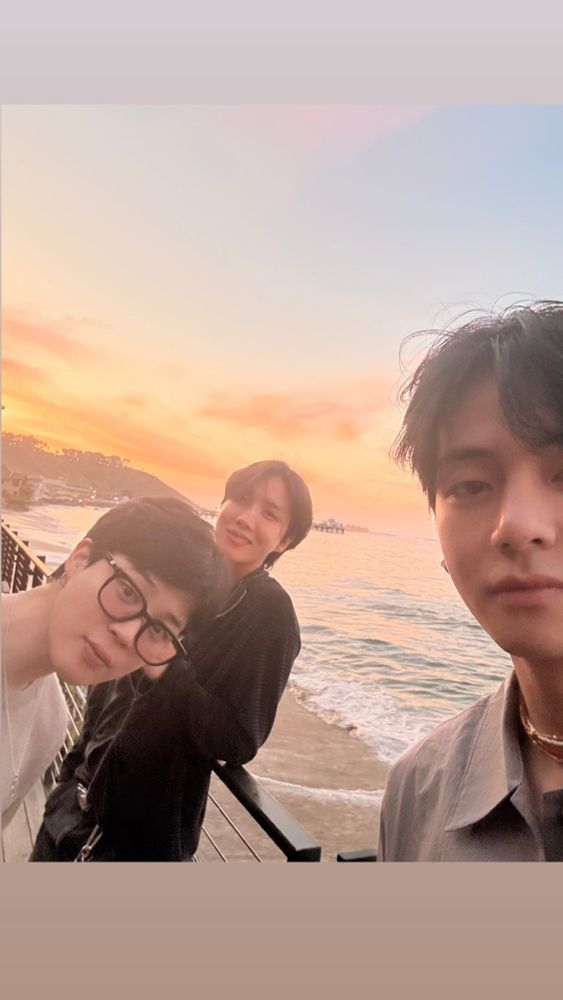 A selca that Taehyung is taking of Jimin and Hobi (V, Jimin, and J-Hope of BTS). The sun is setting, creating a beautiful grayish blue, yellow, and orange gradient on top of the sea. Only half of Tae's face is in the shot, Hobi is leaning against a railing, and Jimin is leaning forward to get into the photo