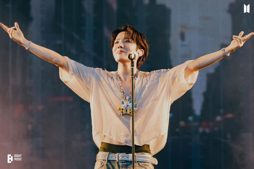 Hobi raising both of his arms on stage as he's standing in front of the mic stand with a solemn look on his face. The stage behind him is pretty dark but Hobi is illuminated beautifully in a yellow-ish organe light from multiple angles. The shadows are blue-ish, creating a nice contrast