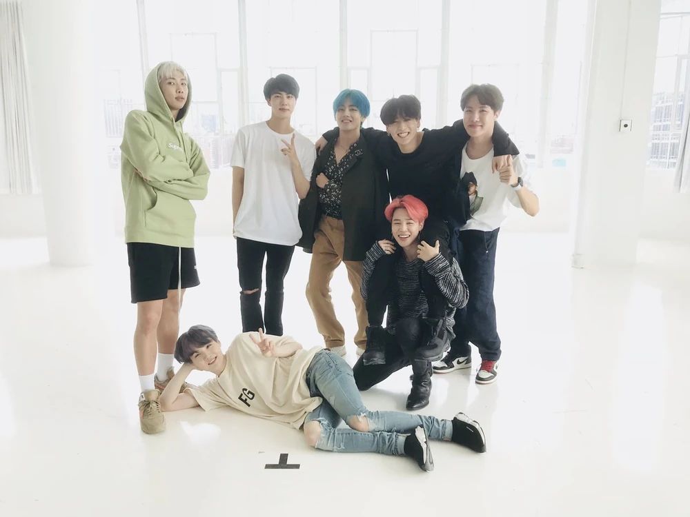 BTS in 2019 in a stark white room after filming a dance practice for their song Boy With Luv. 

Yoongi is laying on the floor in front of them in his famous "draw me like one of your French girls" pose. He's wearing a white FG t-shirt and ripped denim jeans, and his hair is a dark silver. He's smiling, his eyes forming happy crescents.

Namjoon is standing almost right above Yoongi's head with his arms crossed. He's wearing a lime green hoodie with the hood up, and black shorts. His hair is platinum blond. He has a serious look on his face.

Jin is holding up one V sign with a small, barely-there lopsided smile. He's wearing a white t-shirt, and ripped black pants. His hair appears to be black.

Taehyung is smiling while crossing his arms. His hair is an electric blue, and he's wearing a black jacket that's draped across his shoulders, a black button up shirt underneath with white patterns on it, and light brown pants. Jungkook next to him has one arm resting on his shoulders.

Jungkook is riding on Jimin's shoulders, giving a wide, positively delighted, scrunchy smile. His arms are draped across Tae's and Hobi's shoulder for support. He's wearing all black with his hair also being black.

Jimin is kneeling under Jungkook as he's supporting him on his shoulders. He's holding both of Jungkook's legs and is smiling widely. He's wearing a black and white striped shirt and everything else is black. His hair is a peachy pink.

Hobi is supporting Jungkook's other arm. He's holding a thumbs up while giving the camera a close-lipped smile. He's wearing a white t-shirt with a print of a portrait in the center, and dark blue denim jeans. His hair is dark brown.