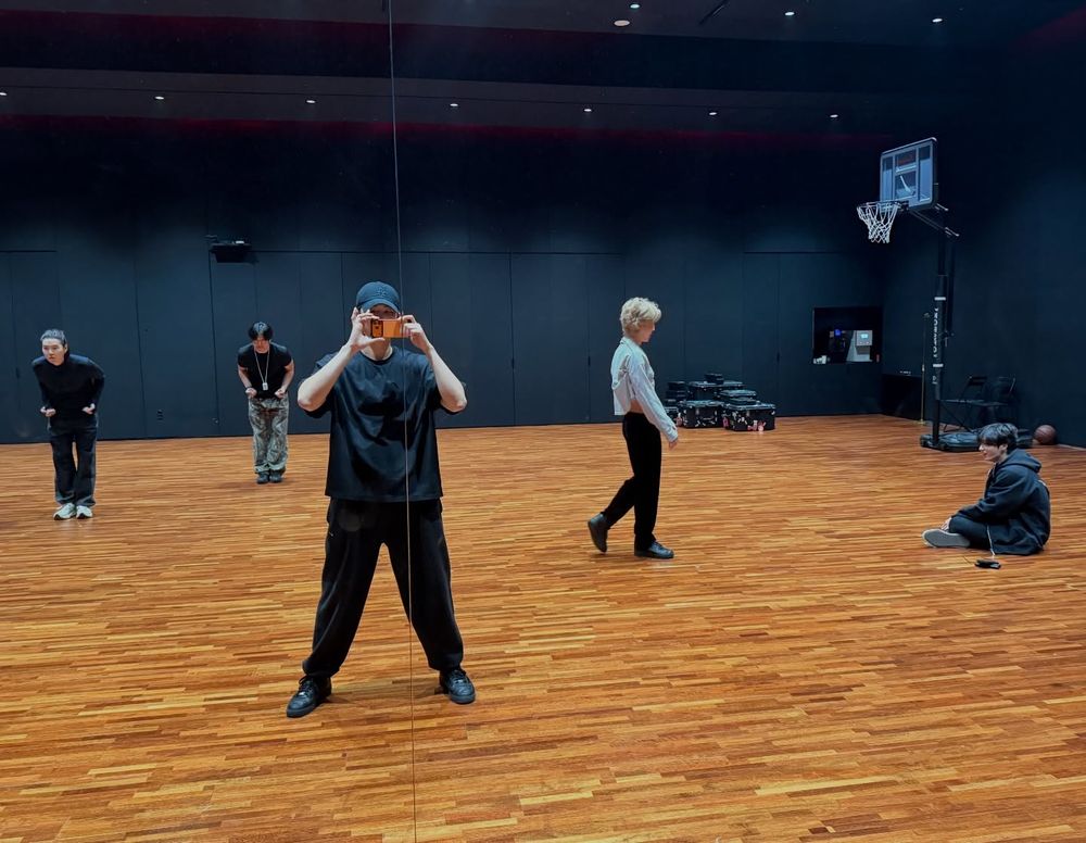 A photo taken by Namjoon of their dance practice room with 4 BTS members in total and 1 staff member. 

Namjoon is standing in front of the mirror and holding up his orange phone. He's wearing a black cap, a black t-shirt, black pants, and black shoes. The phone he's holding up is covering his face.

Yoongi is in the background in the far left, leaning over with his hands slightly above his hips, apparently going through a choreagraphy. His hair is black and tied back. He's also wearing a black pullover, black pants, and white shoes. His features a fairly small so it's hard to tell but he looks like he's concentrating.

A staff member is making the same motions as Yoongi.

Jimin is captured mid-walk as he's walking towards Jungkook. Jimin's hair is blond, and he's wearing white shirt, black pants, and black shoes. The shirt is bunched up under his arm so he's giving us a little peek of his bare midsection.

Jungkook is sitting on the dance floor in the far right. His hair is black, he's wearing a black hoodie, black pants, and the undersides of his shoes are white. He seems to be looking and smiling at something to the far left.