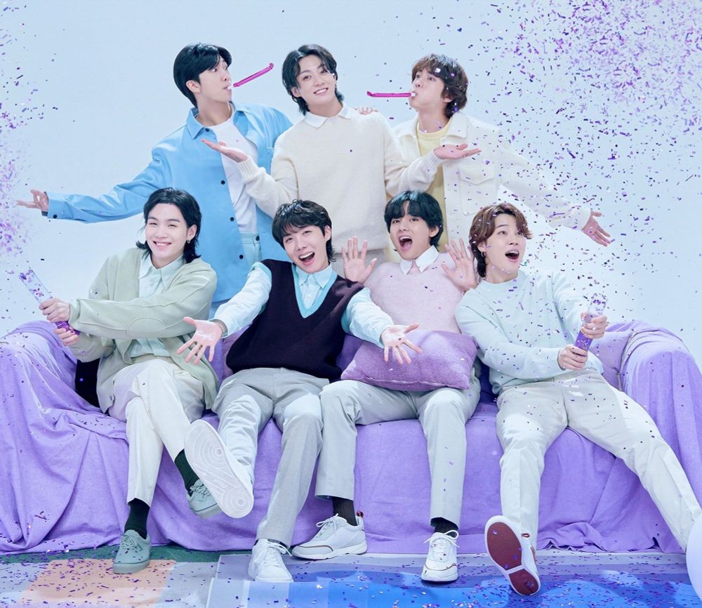 BTS Take Two (2023) era. Yoongi, Hobi, Tae, and Jimin are sitting on a purple couch while Namjoon, Jungkook, and Jin stand behind it. All of them are celebrating with Yoongi and Jimin popping party poppers, and Namjoon and Jin are blowing party noise makers. Everyone (who's mouth isn't occupied) is smiling widely