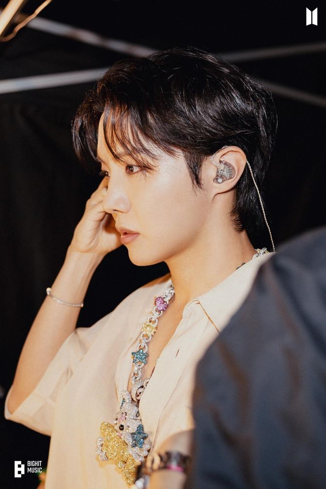 A close up photo of Hobi (J-Hope of BTS) getting ready to perform. He's wearing a loose off-white jacket with nothing underneath, and his sparkly custom-made necklace which is a white cartoony character that's holding big golden "Hobi" letters. Hobi's looking off to the side with a concentrated look