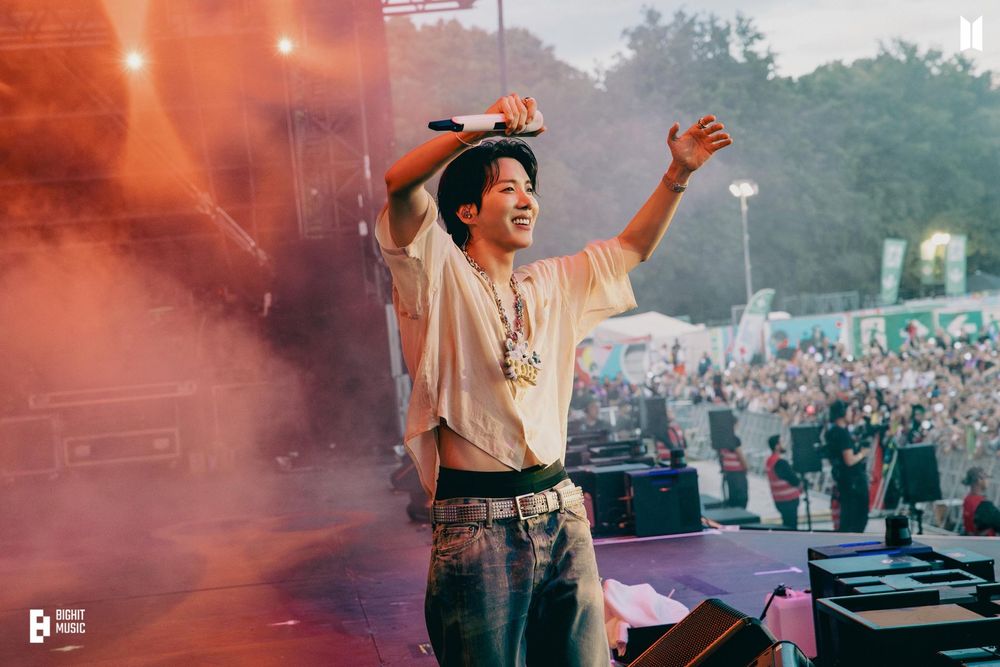 Hobi on stage holding up both arms, wearing the same outfits I described in the last image but here we can also see his blue denim jeans. He's smiling as he's looking at ARMY. We can see half of the stage which has orange red lighting at the moment, and half of ARMY in the background
