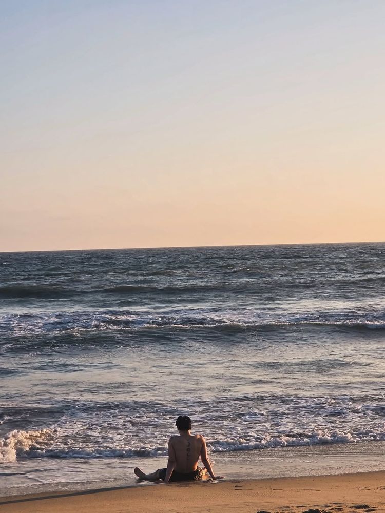 Jimin sitting on the beach in front of the sea with his legs spread wide and his arms behind him to hold up his torso. We immediately can tell it's him because of the moon tattoos on his back. The sun is setting so it's giving the photo a subtle orange glow. 