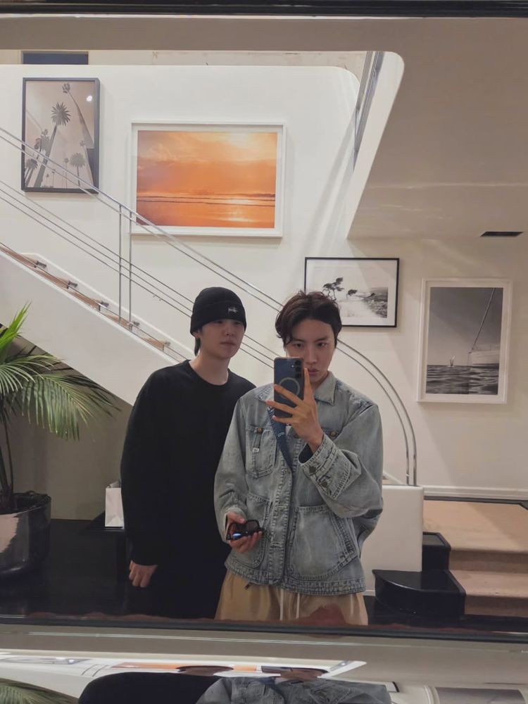 A photo in front of the mirror of Hobi and Yoongi. They're wearing the same outfit as in the first photo I've described so all black for Yoongi and a faded denim jeans jacket for Hobi. Hobi is holding up his phone and looking into it, and holding his sunglasses in the other hand while Yoongi stands beside him and looks into Hobi's phone.

They're in a room that looks like a lobby. There are stairs behind them with a railing, a few landscape photos on the wall, a potted palm tree, and in the front is a shiny counter that reflects Yoongi and Hobi at a weird angle