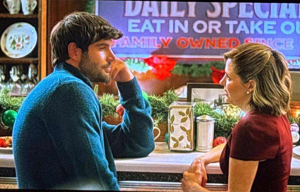 Szene aus "Operation mistletoe", an einem Tresen im Café sitzt links ein Mann-Typ Ken von Barbie, aber mit Bart- mit blauem Pulli, linker Arm auf dem Tresen, die Hand ruht am Kinn, rechts schaut ihm eine junge Frau mit langem Bobschnitt und dunkelrotem kurzärmeligen Kleid in die Augen