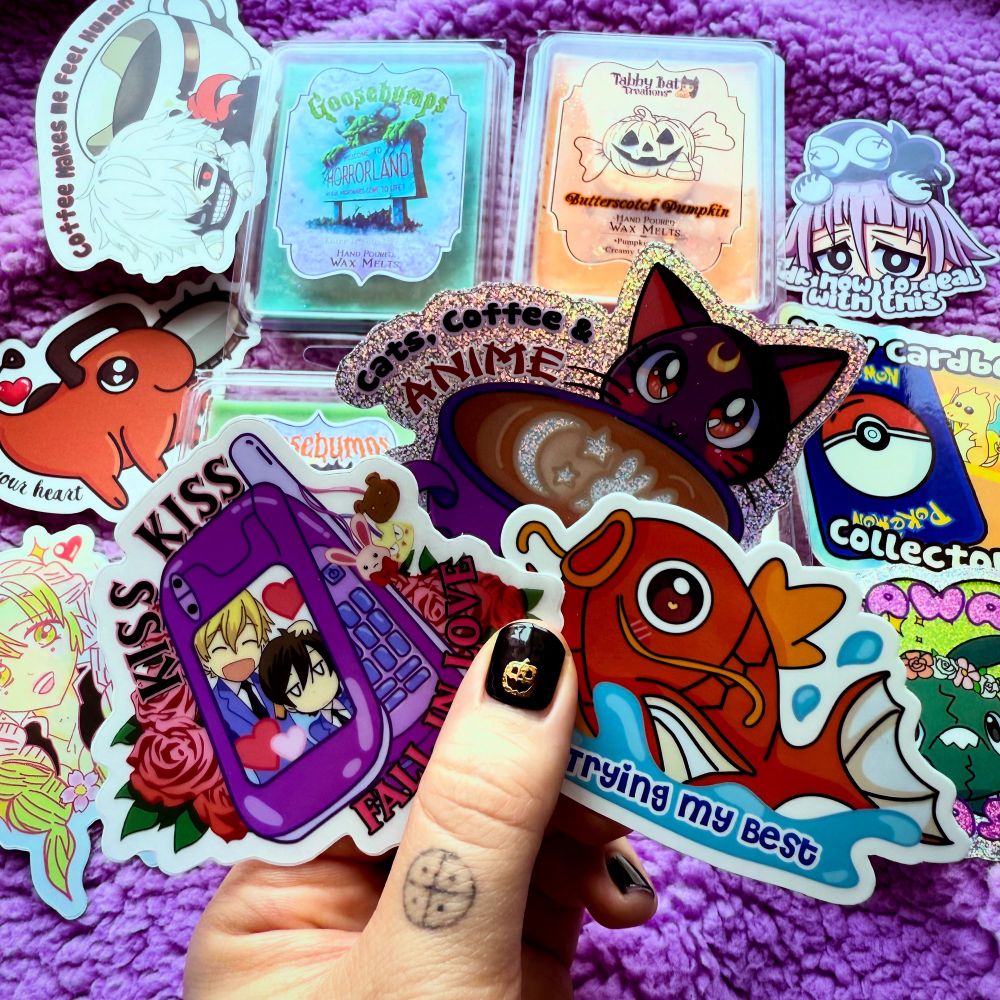 My hand holding stickers from my shop including an Ouran High School Host Club sticker that says “kiss kiss fall in love”, Luna from Sailor Moon with a coffee mug that says “cats, coffee, anime” and a Magikarp sticker that says “trying my best” The background had other fandom stickers and Halloween and Goosebumps themed wax melts