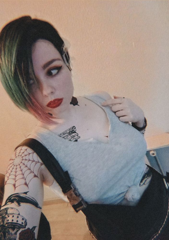 A selfie of a girl dressed as Judy from the videogame Cyberpunk. She has short green pink ombre hair, various tattoos on her arm, a grey knotted tank top and a black dungaree. The background is a cream coloured wall and you can see a chair in one side of the picture in the background 