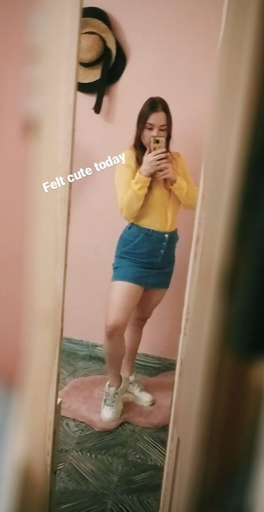 A mirror selfie of a girl standing on a pink irregular rug on top of a gray tiled floor and a pink wall. You can see some hats hanged on the wall and she is wearing a denim button skirt with a yellow shirt. She has brown hair styled down. She's hiding behind her phone and there's a floating text caption saying "felt cute today"