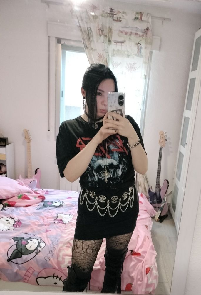 A mirror selfie of the same girl, she has black hair tied up and is wearing a black band tee of the group Slayer, a black pencil skirt with a pentagram chain belt, knee high boots and spiderweb stockings. The background is a white wall with a window, you can see several instruments, like a pink bass and a pink guitar, and there's a pink sanrio bedding on the bed.