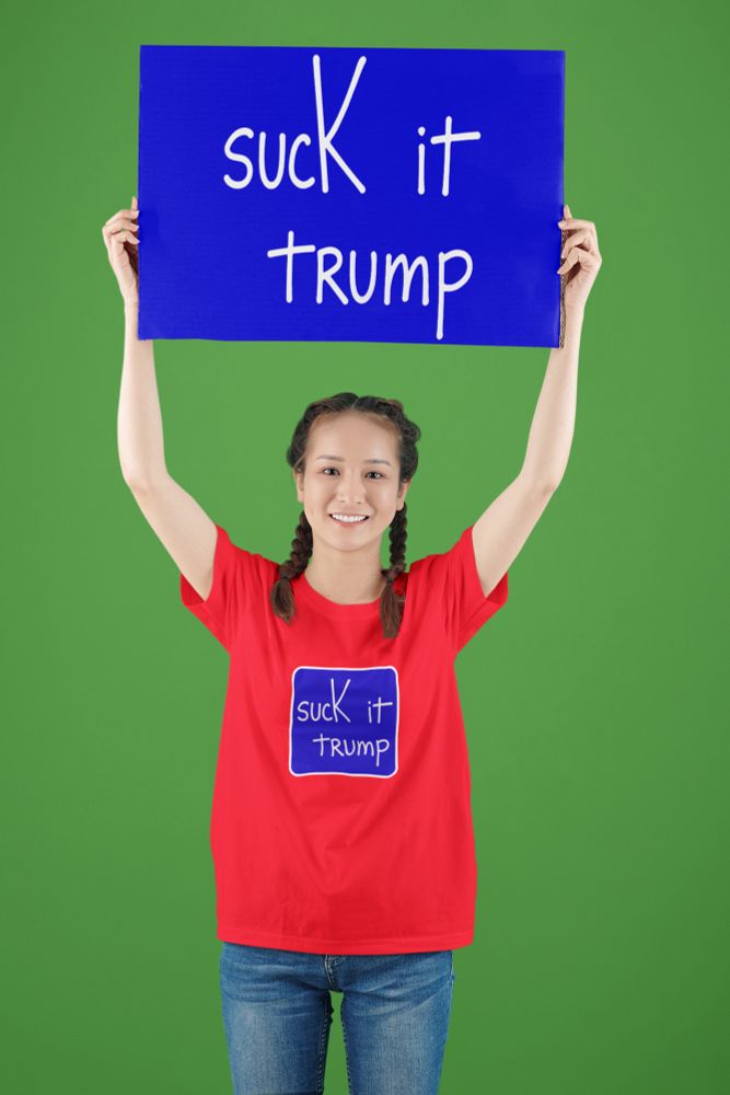A person in a red shirt holds a blue sign overhead. Both the sign and shirt display the same bold text directing an insult at Trump. Green backdrop.