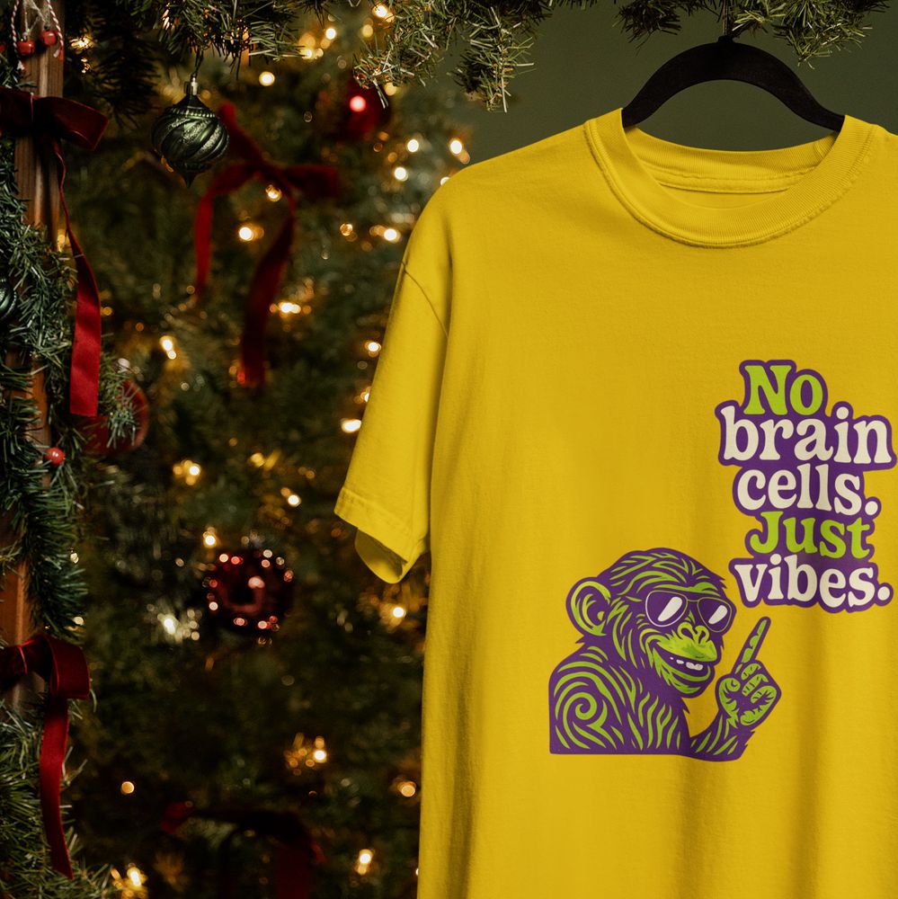 A yellow T-shirt hangs on a black hanger beside a decorated Christmas tree. The shirt features a purple and green graphic of a smiling chimp wearing sunglasses with text that reads No brain cells. Just vibes. Warm holiday lights and ornaments fill the background.