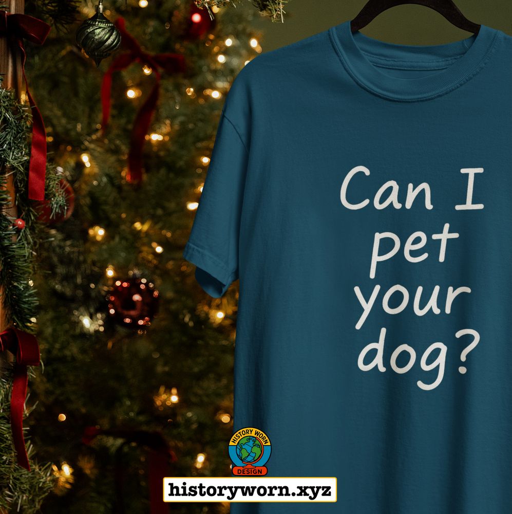 A teal t-shirt with the handwritten text "Can I pet your dog?" hangs in front of a lit Christmas tree. The playful, minimalist design makes it a charming holiday gift for dog lovers. A logo for History Worn Design and the URL historyworn.xyz appear at the bottom. 