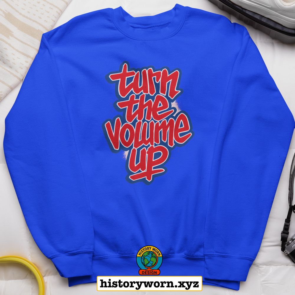 A royal blue sweatshirt with graffiti-style red and white text reading “turn the volume up.” The design has a bold, energetic look. It’s laid out on a bed beside a pair of shoes and a suitcase. At the bottom is a logo and the URL “historyworn.xyz.”