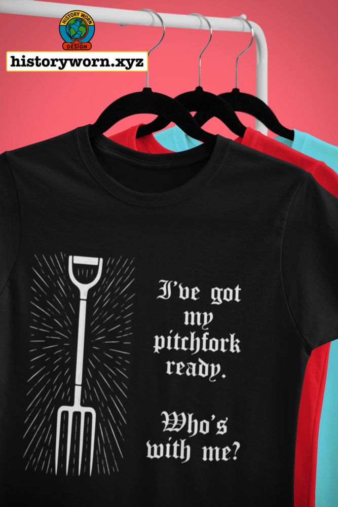 The image shows a black T-shirt on a hanger held by a hand. The shirt features a white graphic of a pitchfork emitting rays, alongside gothic-style text that reads: “I’ve got my pitchfork ready. Who’s with me?”