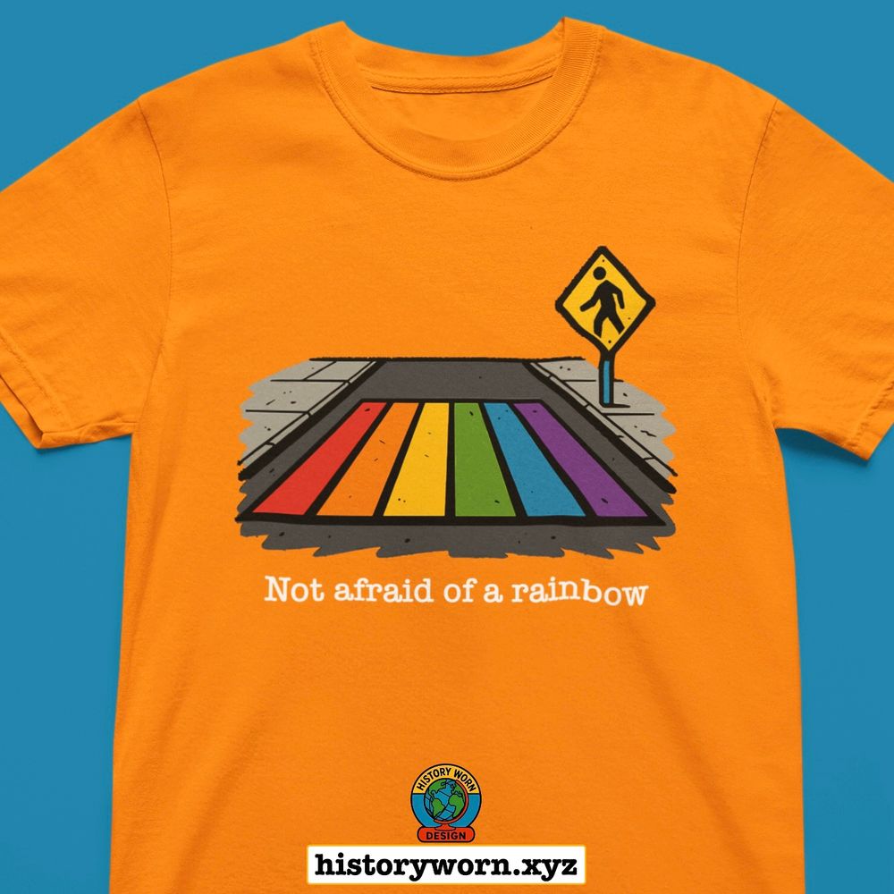 Orange shirt with an illustration of a rainbow painted crosswalk beside a pedestrian sign. Text reads “Not afraid of a rainbow.” A small circular logo appears near the bottom along with the URL historyworn.xyz.