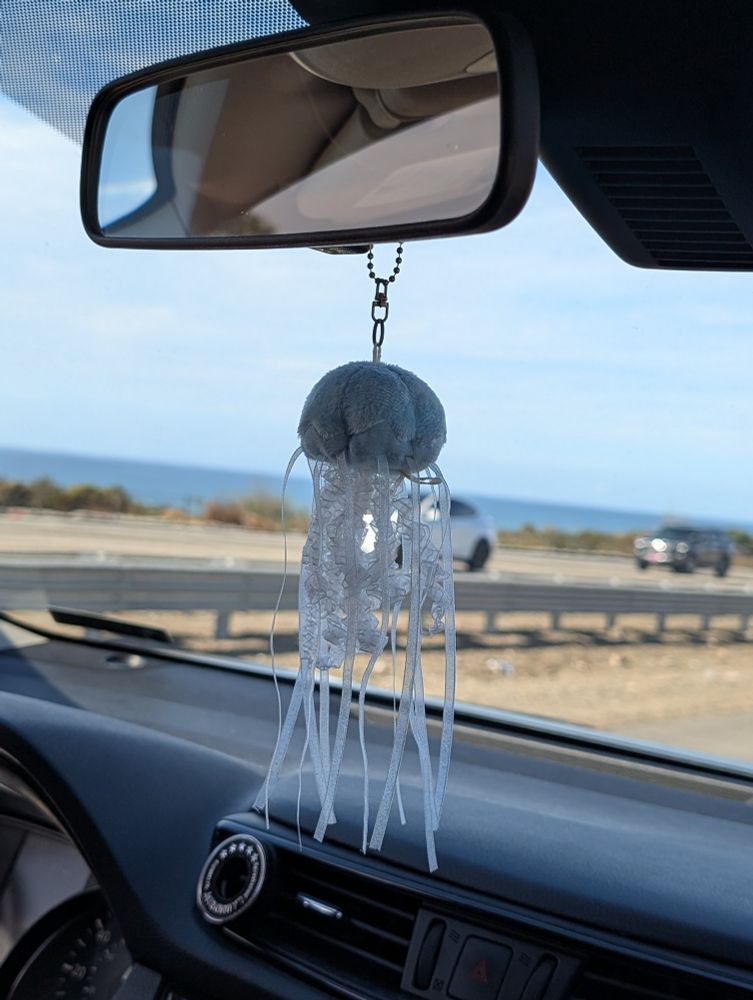 A small, light blue, stuffed jellyfish hangs from  a rear view mirror, which is reflecting the gray roof of the vehicle. The dashboard is also gray. Through the windshield, one can make out the dirt median of the road with a guard rail, two vehicles going the opposing direction, and in the distance is the ocean. 