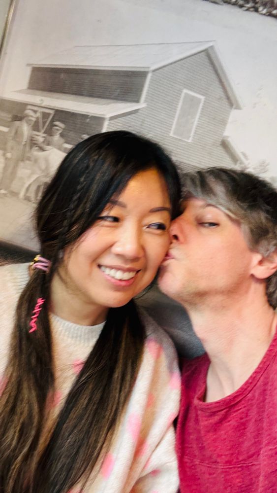 A pretty Asian girl with pigtails is kissed by a scruffy white boy. 