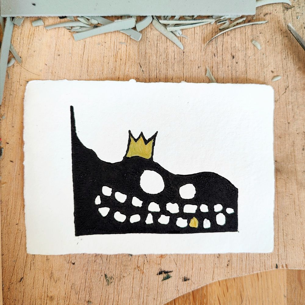 Photo of a linocut print on a wood background, surrounded by strips of offcuts of lino. The print is a grinning pile of black ooze wearing a king hat. The king hat is an unpleasant yellow and the ooze has a gold tooth.