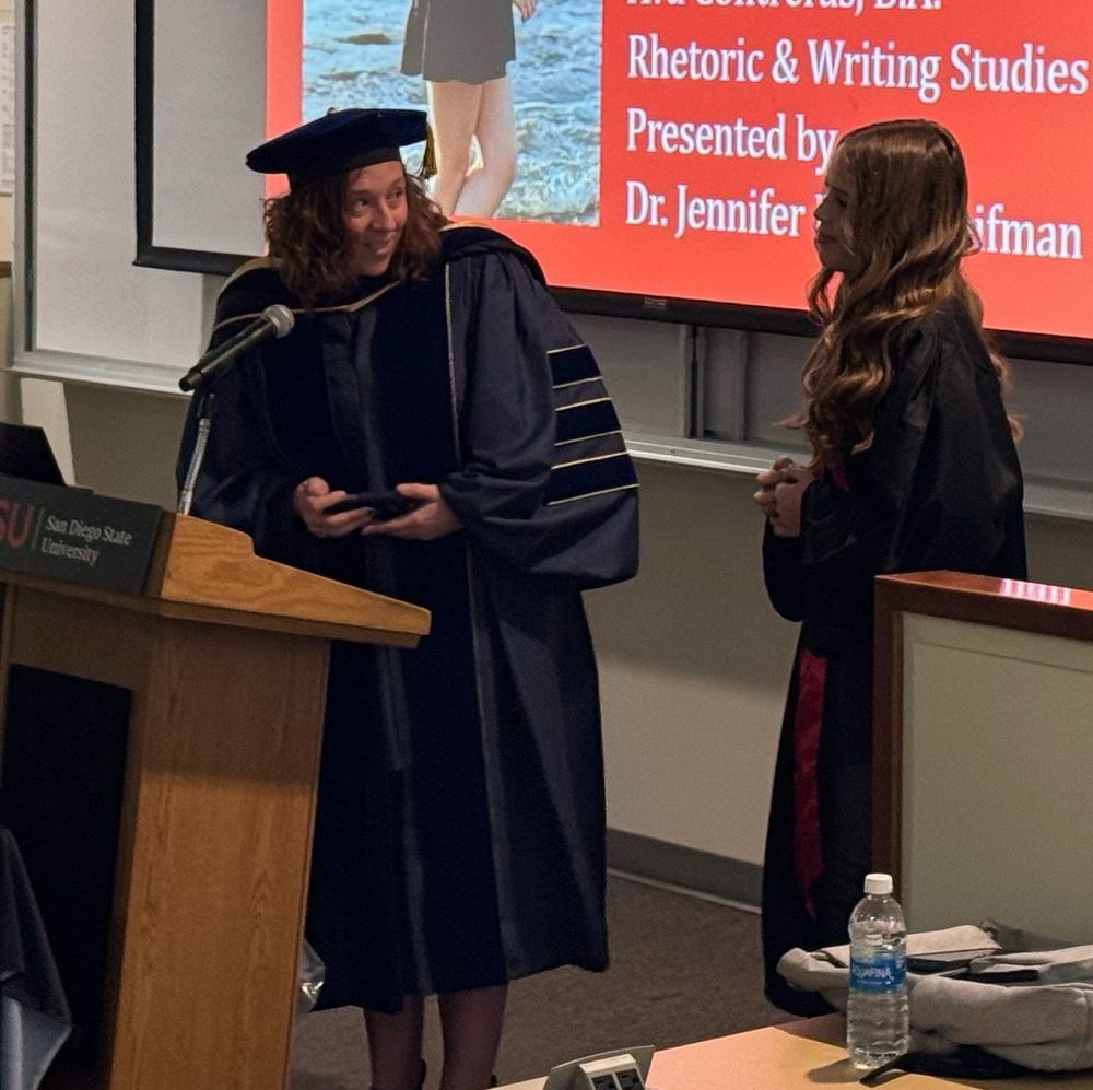 Jennifer stands in a podium in graduation regalia, speaking to the student beside her who is also in a cap and gown. 
