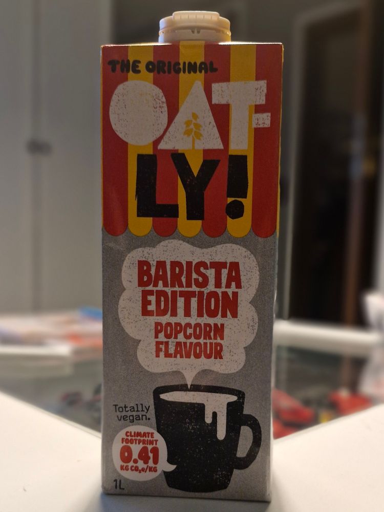 Ein 1-Liter-Getränkekarton der Marke Oatly mit der Aufschrift „The Original Oatly! Barista Edition Popcorn Flavour“. Der Karton ist gelb-rot gestreift, trägt eine schwarze Tasse als Motiv und den Hinweis „Totally vegan“.