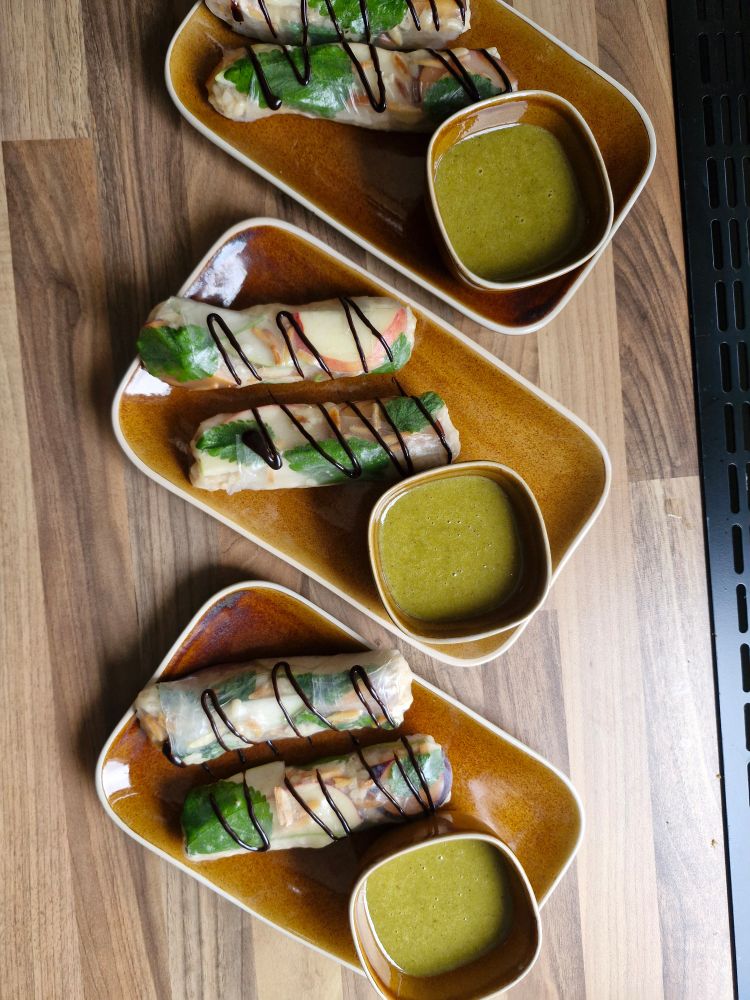 Drei braune rechteckige Teller auf Holzoberfläche, jeweils mit zwei Rollen aus Reispapier, gefüllt mit Obst, Michreis und Zitronenmelisse. Die Rollen sind mit etwas Schokolade verziert. Daneben steht pro Teller eine kleine Schale mit grünem Dip.