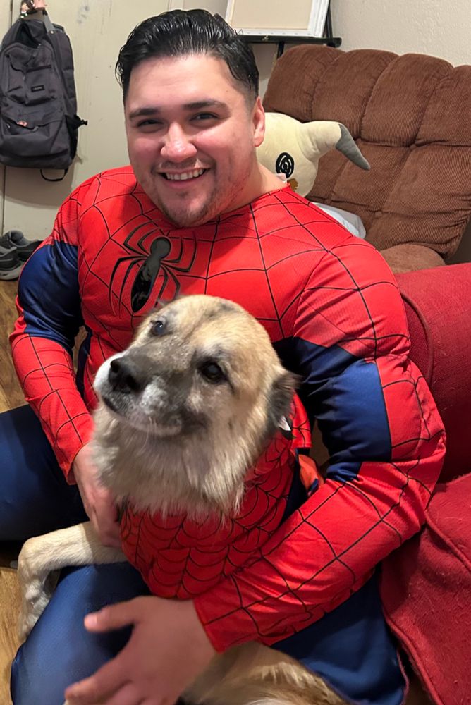 A chubby Latino man with short dark hair and stubble posing with a light brown husky dog, both dressed as Spider-Man