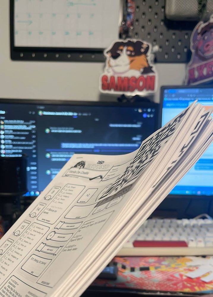 A stack of papers in front of 2 monitors. They are dnd character sheets