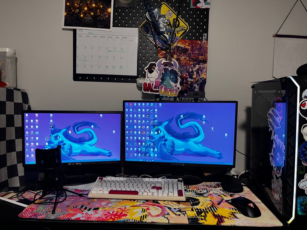 A pc set up with two monitors and a tower. The background has a picture of my fursona. Above the desk is a pin board with furry art and a calendar