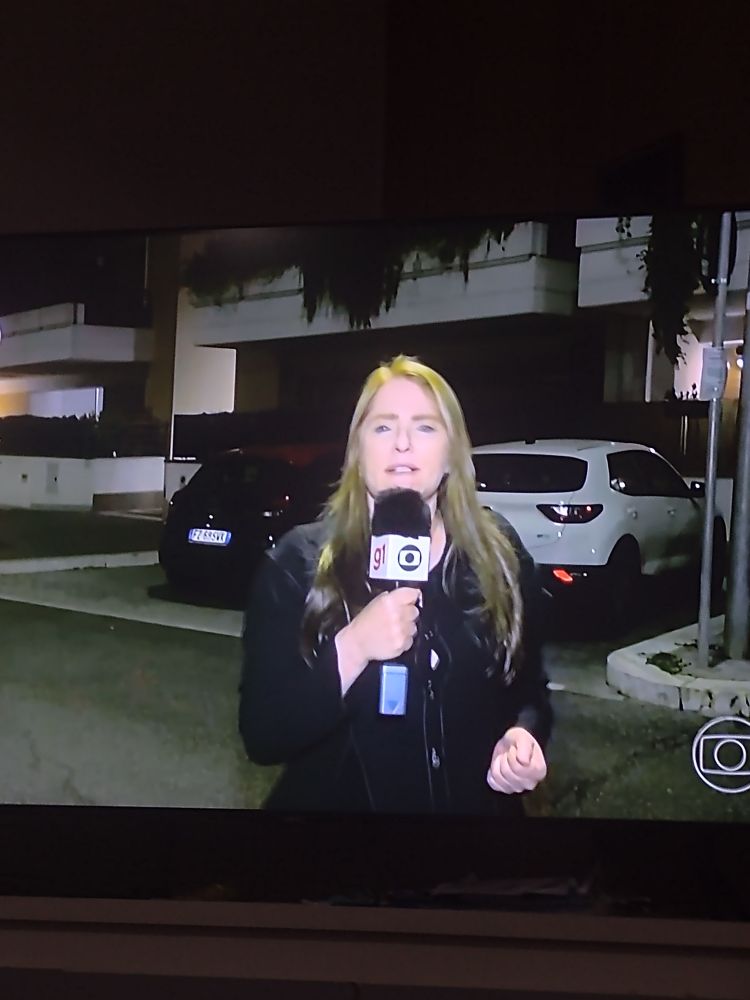 Foto do Jornal Nacional em que a repórter Ilze Scamparini aparece em um prédio diferente do que ela costuma aparece. Está usando um casaco preto e segura no microfone tradicional da TV Globo.