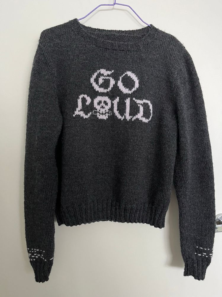 A charcoal-grey crew neck sweater, with the words Go Loud in gothic font on the front upper centre. The sleeves have Morse code embroidery at the wrists, spelling out the phrases “life is too short” and “love is too long”, with a row of three embroidered hearts beneath each. 