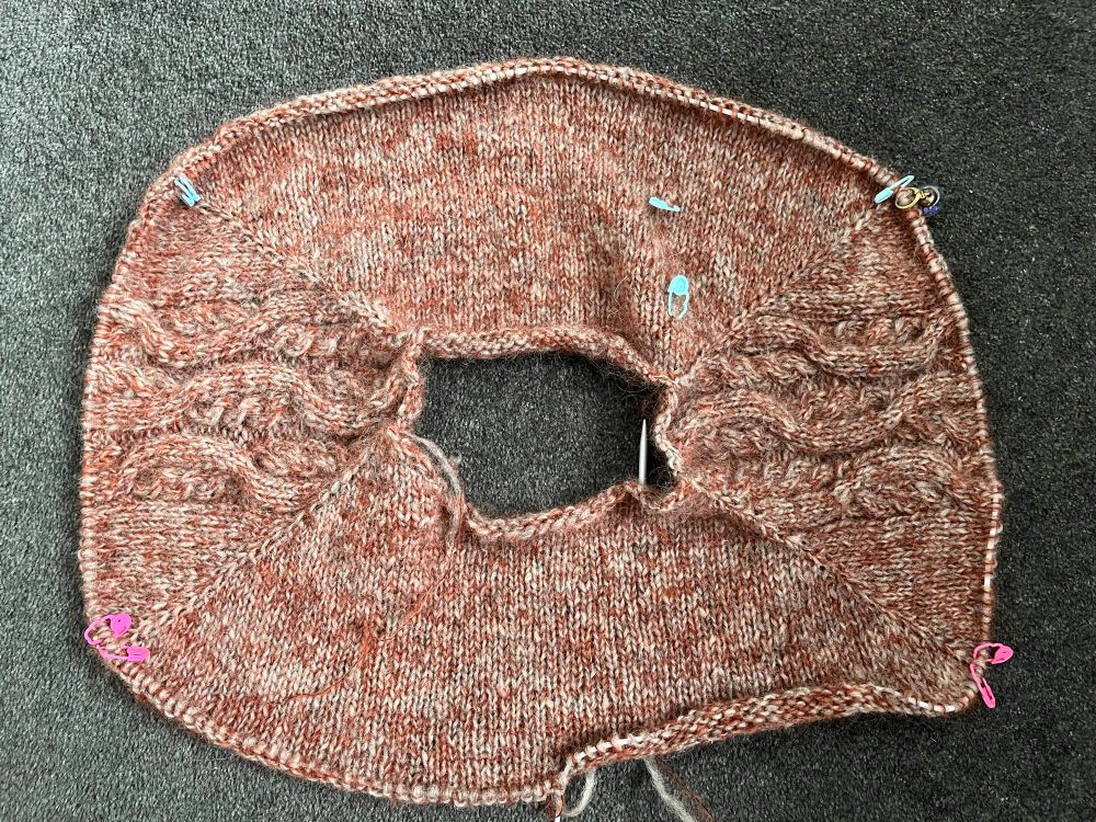 The yoke of a knitted sweater, in a marled rust colour, achieved by holding one stand of dark red-brown alpaca lace yarn with one strand of grey blend unspun. It looks incredibly soft to the touch (and is). There are an obscene number of stitch markers attached to the project, for raglan increases, cable row progress, and other things your maker has already forgotten the moment she stopped looking at the pattern.