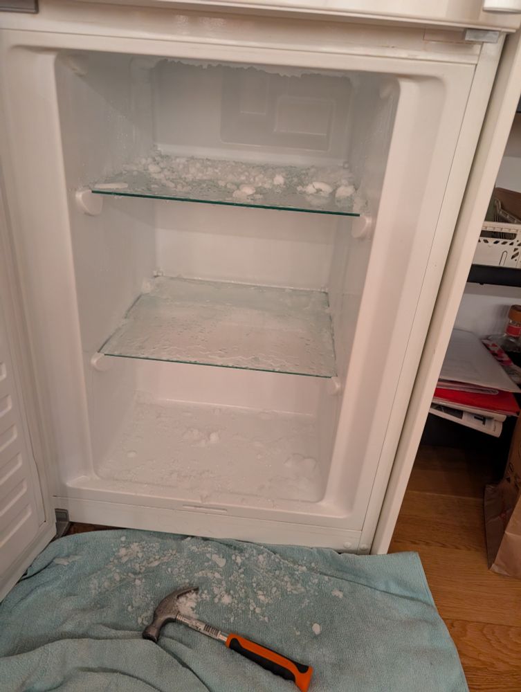 En tom frys med öppen dörr. På golvet framför ligger en hammare på en ljusblå handduk