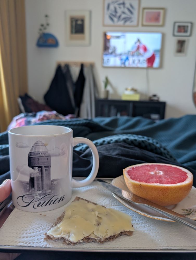 En bricka med kaffe, knäckebröd, grapefrukt i en säng. I bakgrunden står en TV på och visar längdskidåkning 