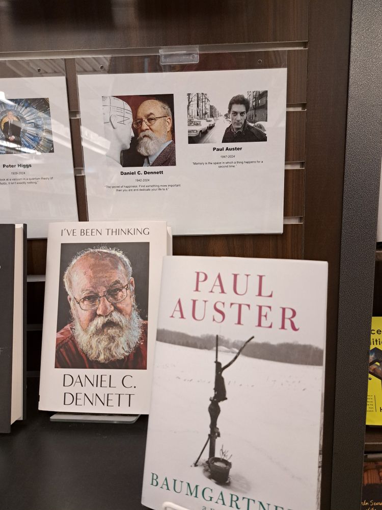 A display of hardcover books shows Baumgartner by Paul Auster and I've Been Thinking by Daniel C Dennett, with Dennett's picture on the cover. The white cover of Baumgartner shows a water pump and frozen bucket of water in a snow-covered field. A placard with photos and the aforementioned quotes from each author is above. Dennett is looking at the camera sideways while facing the head of a white dummy inches away, the dummy's head marked into black squares, one square says the selfish propensity, another square says perception. Auster, 1947-2024 is in tge middle of a street, possibly in New York, looking at the camera.