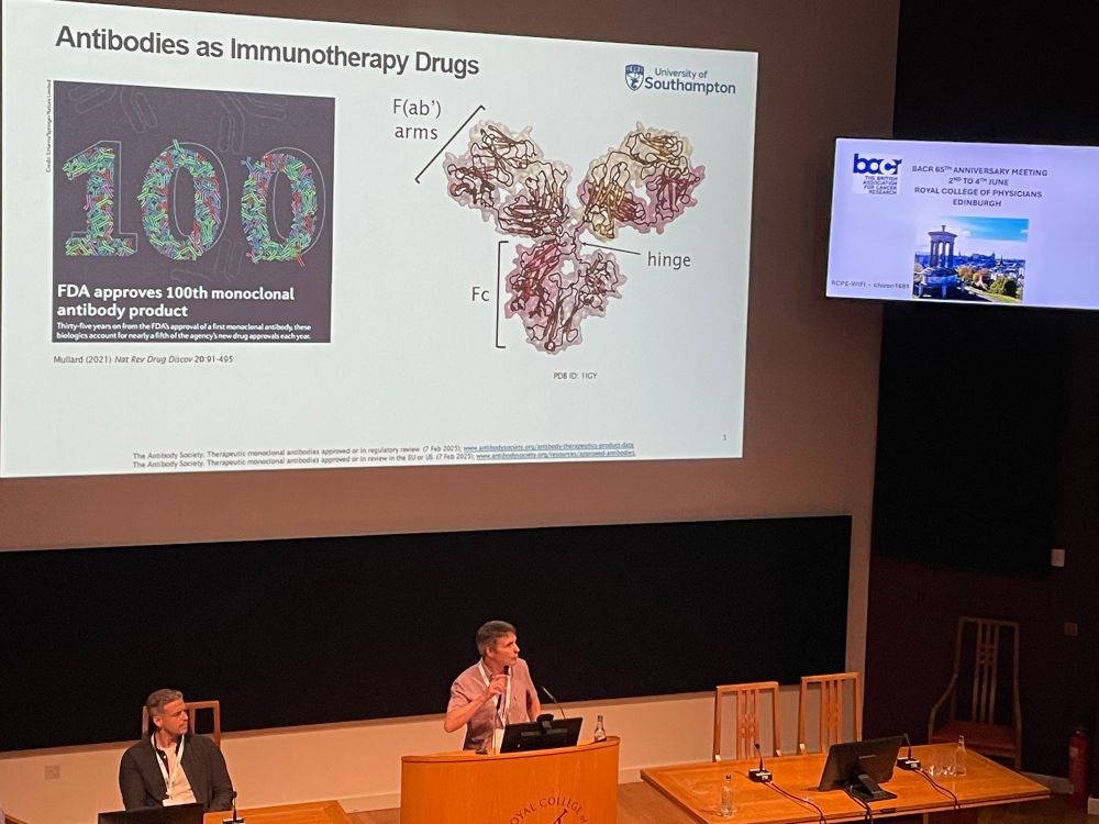 Mark Cragg in a checked short-sleeved shirt is now behind the lectern. Behind and above him is a huge crystal structure of an antibody, and a graphic celebrating the recent FDA approval of the 100th antibody-based drug.