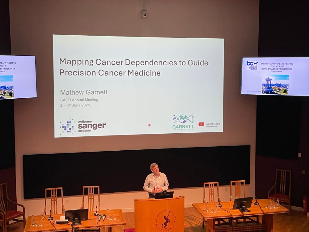 Mathew Garnett stands behind the wooden lectern in a blue shirt. Above him the screen introduces his talk on “Mapping Cancer Dependencies to Guide Precision Cancer Medicine”.