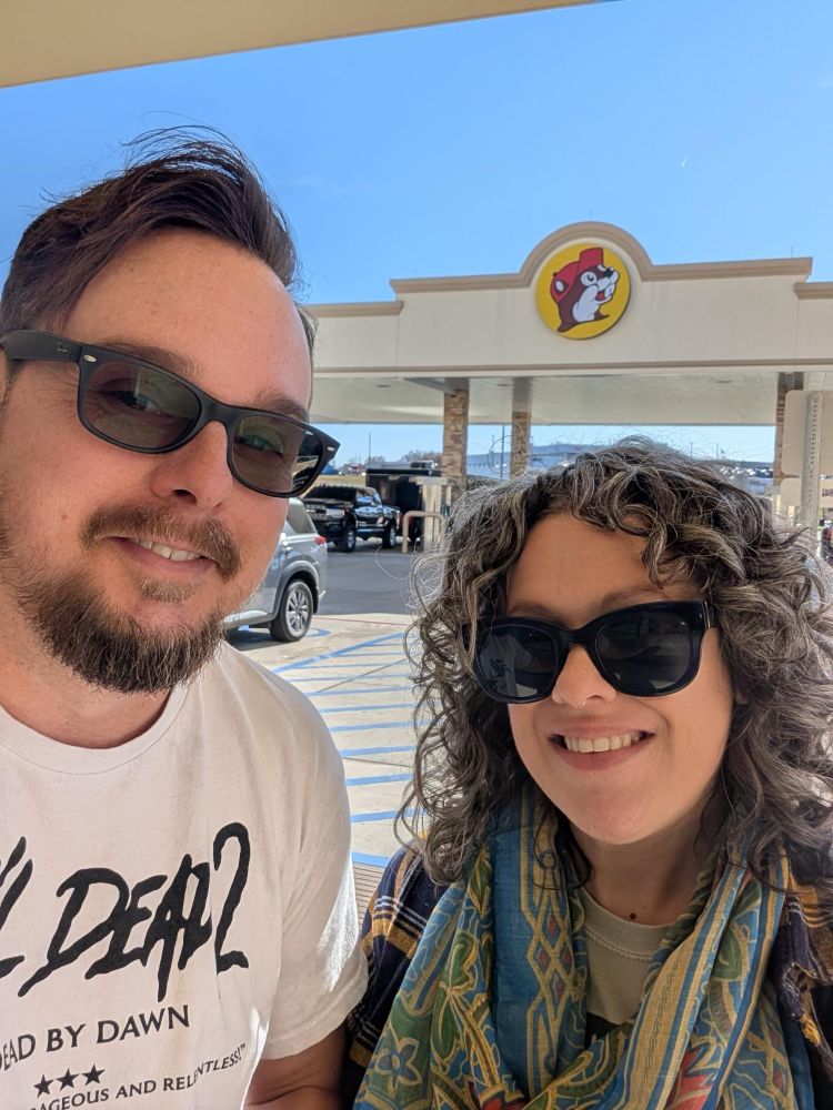 A selfie of a dark haired man in sunglasses (left) and dark haired woman in sunglasses smiling in front of the Buc-ees sign