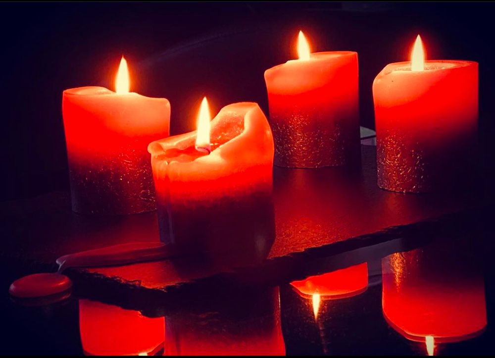 Vier rote, brennende Kerzen auf einer Schiefertafel. Der Hintergrund ist dunkel. Von einer Kerze ist der Wachs auf die Schiefertafel und weiter auf den Tisch gelaufen.