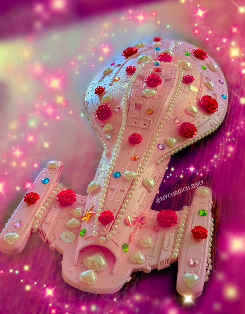 Star Trek Voyager model painted pink covered in heart shaped pearl stickers, rainbow jewel stickers, rose stickers and string of pearl stickers. The background is blurred with added in sparkly pink stars. 

Watermark: @mychadich.bsky