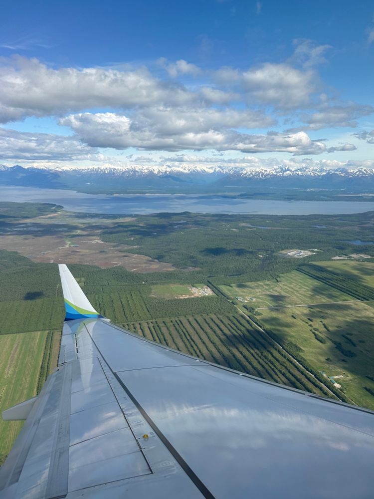 View from the plane arriving into Anchorage. The white wing of an Alaska Airlines plane covers the bottom third of the photo. Beyond the wing is a lush green permafrost field with the ocean and tall, snow capped mountains in the distance. The sky is bright blue with puffy clouds. 