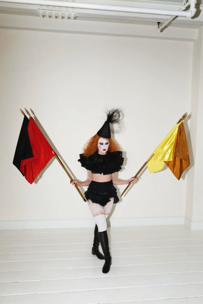 Chappell Roan looking like a Runescape character in a white room dressed in a little black Pierrot outfit holding two flags at her side 