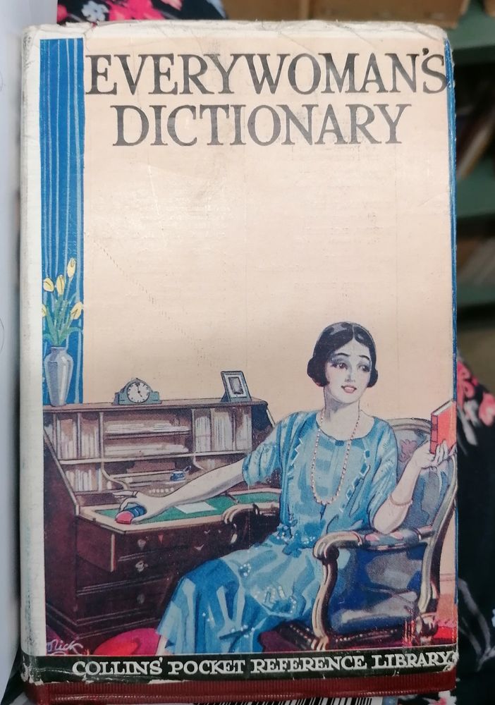 The cover of Everywoman's Dictionary from 1928, featuring an elegant woman sitting at a writing desk. She is wearing a blue drop-waisted dress which one must assume has no pockets. 