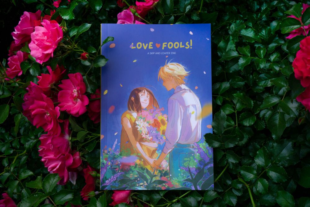 The image shows LOVE FOOLS! A Skip and Loafer Zine, set against a background of leaves and flowers. The cover features an illustration of Mitsumi and Shima facing each other, holding hands in a patch full of flowers. Mitsumi is smiling and Shima is turned towards her, his expression not visible to the viewer though there is a blush on his cheeks. 