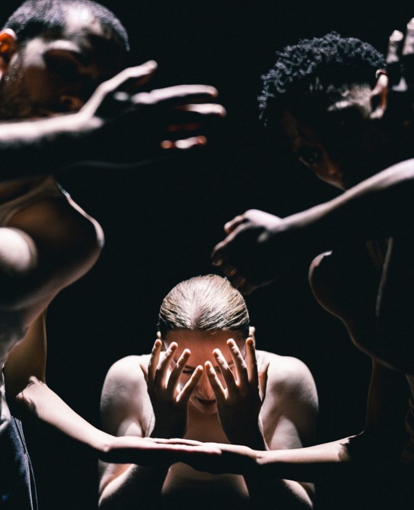 Image from Figures in Extinction - a spot lit woman covers her face as hands and arms surround her