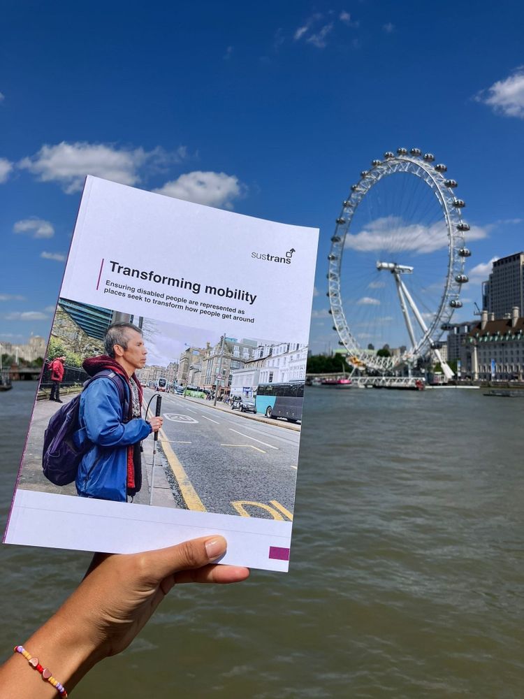 A hand holding the Transforming Mobility report in front of the London eye. The river Thames, buildings and a blue sky can also be seen