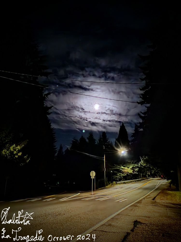 A photo of a crossroads at night with the moon looming overhead through some clouds and a street lamp.