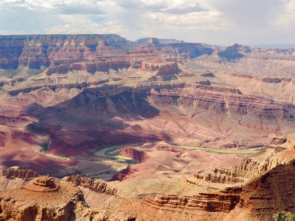 The canyon looks less deep here with the Colorado River clearly seen meandering through it. Tans, browns, and reds make up the landscape. 